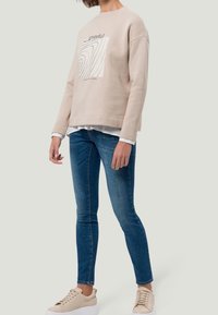 Beige sweatshirt met grafisch ontwerp, gelaagd over een wit shirt; gecombineerd met blauwe jeans en beige sneakers. Casual, relaxte pasvorm.