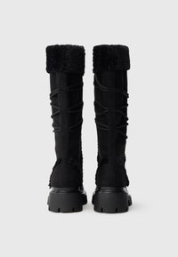 Cizme negre până la genunchi, cu manșetă moale din blană artificială, material din piele întoarsă, detaliu cu șireturi pe partea din spate și talpă texturată din cauciuc.