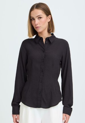 Mujer con cabello liso hasta los hombros, viste una camisa negra ajustada con botones y pantalones blancos, sobre un fondo claro y sencillo.