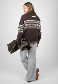 Femme de profil portant un pull marron à motifs, un pantalon large gris, tenant une veste sombre pliée, avec des chaussures marron clair sur un fond uni.