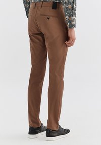 Brązowe bawełniane spodnie w slim fit, z tylną kieszenią zapinaną na guzik, w połączeniu z czarnymi sneakerami. Gładka tekstura i elegancki design.