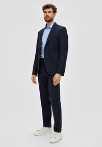 Marineblauer Anzug mit taillierter Passform, bestehend aus einem Zwei-Knopf-Jacket und passender Hose, kombiniert mit einem hellblauen Hemd und weißen Sneakers.