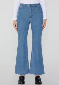 Vaqueros de mezclilla azules de cintura alta con campana, con un detalle de corte en el dobladillo y costuras en contraste. Combinados con zapatos negros.