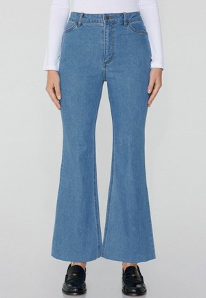 Vaqueros de mezclilla azules de cintura alta con campana, con un detalle de corte en el dobladillo y costuras en contraste. Combinados con zapatos negros.