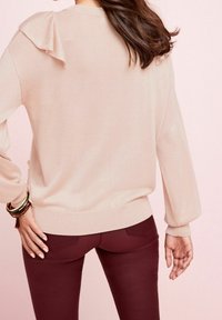 Hellrosa gestrickter Pullover mit Rüschendetail an der Schulter, kombiniert mit schmal geschnittenen, dunkelburgunderfarbenen Hosen. Weiche Textur, lässiges Design.