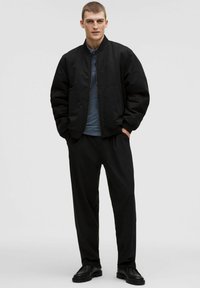 Młody mężczyzna stoi z rękami w kieszeniach, ubrany w czarną kurtkę bomberkę, niebieską koszulę, czarne spodnie i czarne buty na jednolitym tle.