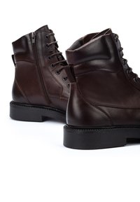 Botas de tobillo de cuero marrón con un cierre de cordones frontal y cremallera lateral, parte superior texturizada y suela de goma negra con detalle de costura.