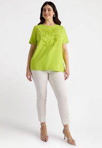 Limoengroene top met korte mouwen en bloemenborduursel. Gecombineerd met witte slim-fit enkelbroeken en zilveren bandjeshakschoenen.