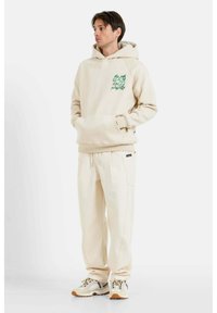 Felpa beige con grafica verde sul lato sinistro del petto, abbinata a pantaloni jogger beige coordinati. Entrambi gli articoli presentano una texture morbida e una vestibilità rilassata.