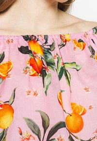 Donna che indossa un top rosa a spalle scoperte con una stampa di frutta arancione brillante e foglie verdi, che mette in evidenza la clavicola e un orecchino a cerchio dorato.