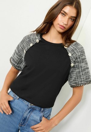 Schwarzes kurzärmliges Oberteil mit Ärmeln im Tweed-Muster und Knopfdetails. Das Shirt ist tailliert und wird mit hellblauen Jeans kombiniert.