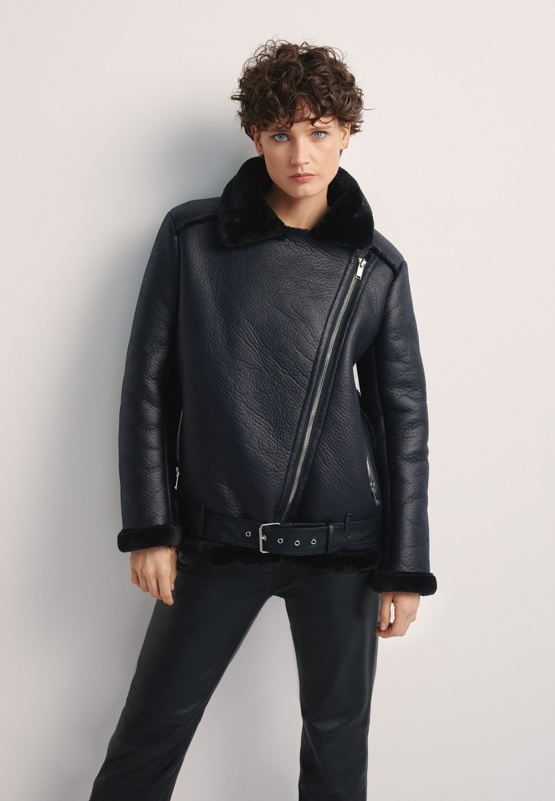 Czarna skórzana kurtka o teksturowanym wykończeniu, z futrzanym kołnierzem, asymetrycznym zamkiem błyskawicznym i paskiem w talii. Nosi się ją na czarnych spodniach.