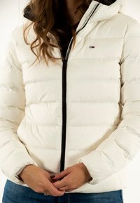 Chaqueta acolchada blanca con costuras horizontales, cremallera negra y capucha. Presenta un pequeño logo en el pecho, textura suave y diseño aislante.