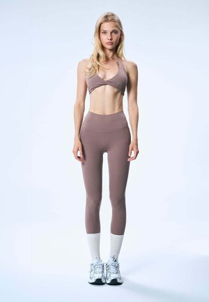 Młoda kobieta stoi ubrana w mauve sportowy biustonosz i legginsy, białe skarpetki i srebrne trampki na jednolitym jasnym tle.