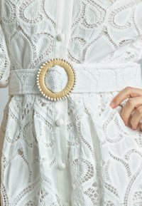 Biała koronkowa sukienka z zawiłymi wzorami i teksturowaną powierzchnią, przewiązana w talii szerokim paskiem z okrągłą, plecioną klamrą.