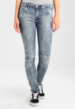 Vaqueros ajustados de mezclilla de color azul claro, desgastados, con parches en las rodillas y detalles de desgaste, combinados con zapatillas negras y blancas.