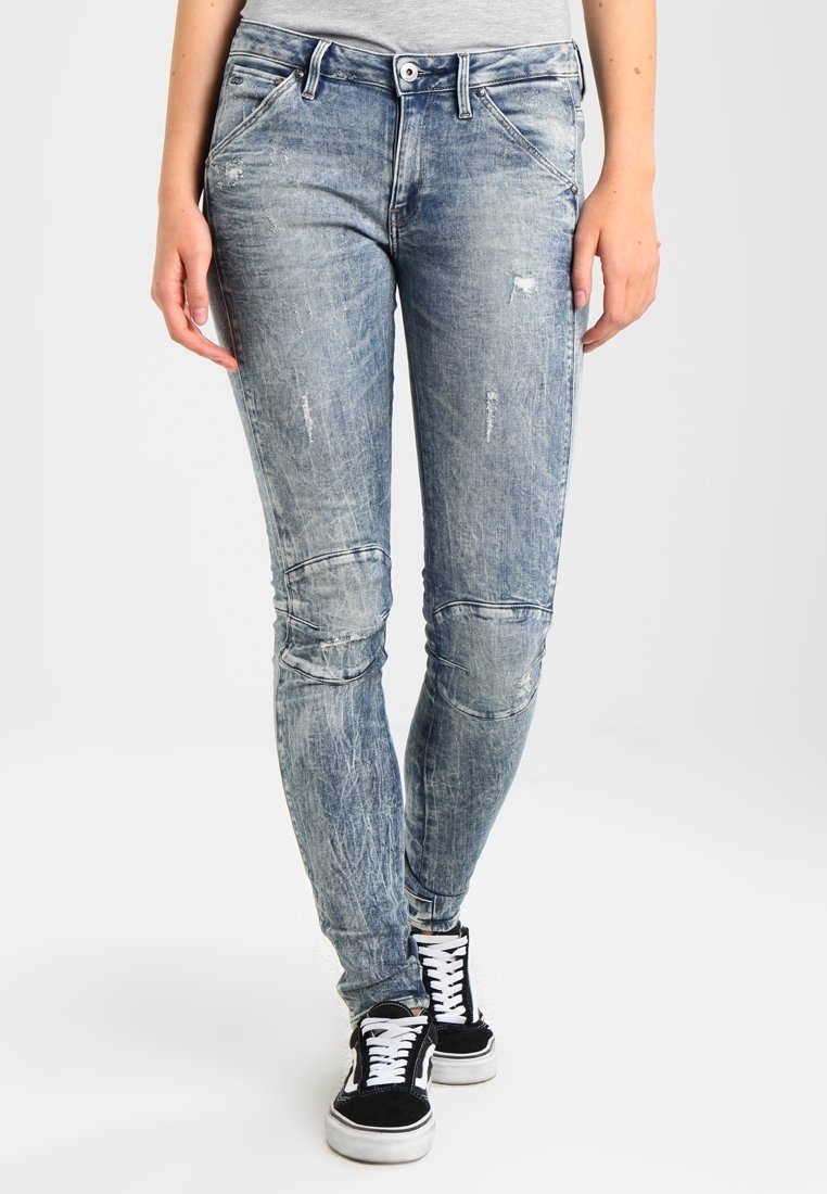 Vaqueros ajustados de mezclilla de color azul claro, desgastados, con parches en las rodillas y detalles de desgaste, combinados con zapatillas negras y blancas.