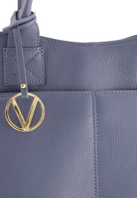 Leather tote bag in a muted blue color featuring a textured finish. Accented with a gold circular logo charm and contrasting stitching.