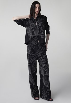 Femme portant un ensemble assorti noir avec de grands motifs floraux blancs en contour, debout avec une main derrière la nuque, sur un fond uni.