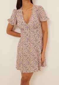 Jumpsuit floral dengan garis leher V yang dalam, lengan pendek puff, dan kain ringan dengan campuran pola ungu, kuning, dan hijau.