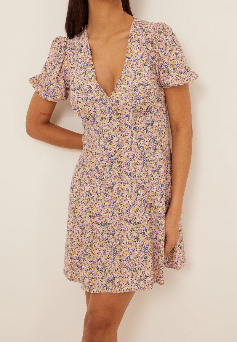 Jumpsuit floral dengan garis leher V yang dalam, lengan pendek puff, dan kain ringan dengan campuran pola ungu, kuning, dan hijau.