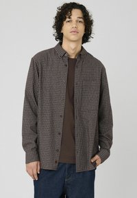 Chemise à carreaux marron et crème, avec un col et une fermeture à boutons sur le devant, ainsi qu'une poche poitrine unique, portée par-dessus un t-shirt marron. Pantalon en denim.