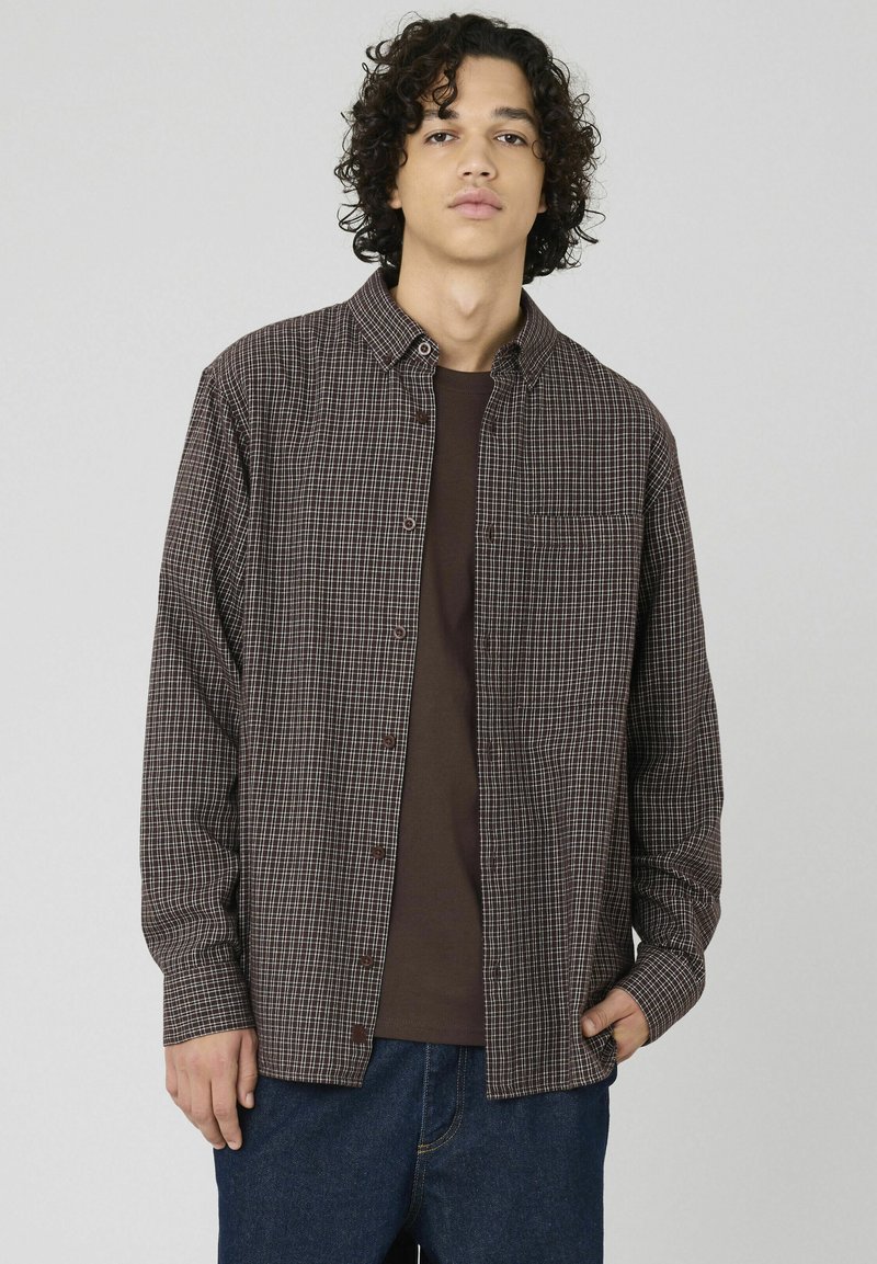 Chemise à carreaux marron et crème, avec un col et une fermeture à boutons sur le devant, ainsi qu'une poche poitrine unique, portée par-dessus un t-shirt marron. Pantalon en denim.