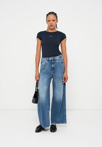 T-shirt ajustado azul-marinho, calças de ganga de corte largo em azul claro com bainha desfiada e mala preta. Tecido suave e ligeiro elasticidade na t-shirt.