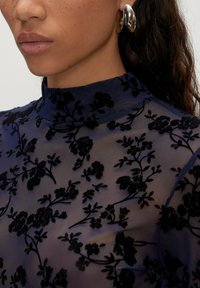 Durchsichtige marineblaue Bluse mit hohem Kragen, die mit schwarzem Blumenstickerei, zarter Textur und eleganten Mustern verziert ist. Silberner Ohrring sichtbar.