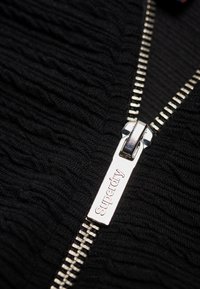 Tirette de fermeture éclair en métal gravée du mot « Superdry » sur un tissu noir texturé.