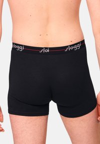 Schwarze Baumwoll-Boxershorts mit rotem elastischem Bund, der mit "sloggi"-Branding versehen ist. Glatte Textur, körperbetonter Schnitt und Länge bis zur Mitte des Oberschenkels.