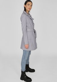 Lichtgrijze trenchcoat met een ceintuur in de taille, notch kraag en voorzakken, gecombineerd met lichtblauwe jeans en zwarte enkelboots.