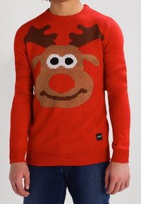 Pull rouge en tricot avec un grand visage de renne, comprenant des bois marron, des yeux blancs et un nez rouge. Poignets et ourlet côtelés.