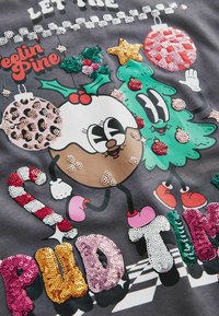 Grafisches T-Shirt mit einer Lebkuchenfigur und einem Weihnachtsbaum, verziert mit bunten Pailletten, auf einem dunkelgrauen Stoffhintergrund.