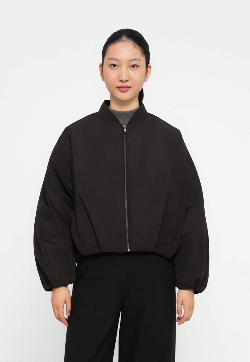 Zwarte cropped bomberjack met een ritssluiting, oversized mouwen en een hoge kraag. Het materiaal lijkt glad en heeft een lichte textuur.