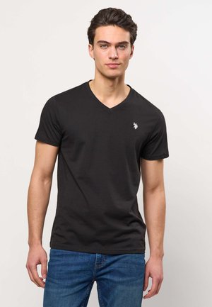 Young man with short dark hair, wearing a black V-neck T-shirt with a small white logo and blue jeans, standing against a plain background.