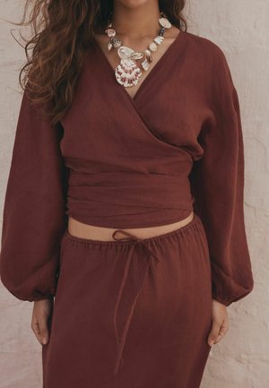 Kvinde iført et dybrødt wrap-top- og nederdelssæt med en skalsmykke og perlekæde, stående mod en tekstureret lys væg.