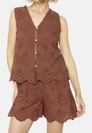 Haut en dentelle œillet marron sans manches avec boutons et short assorti aux bords festonnés, porté par une personne debout avec une main vers le bas.