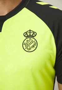 Maglia sportiva giallo-verde con dettagli neri. Presenta un collo rotondo e un logo stampato con una corona. Pattern in tessuto testurizzato visibile.