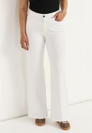 Person iført hvide højtaljede vide bukser, sort top, brune sneakers, med den ene hånd i lommen og den anden afslappet ved siden.