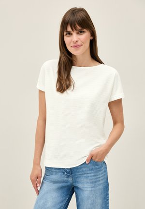 Haut blanc texturé avec un col rond et des manches courtes, assorti à un jean bleu clair. Le tissu présente un motif ondulé subtil.