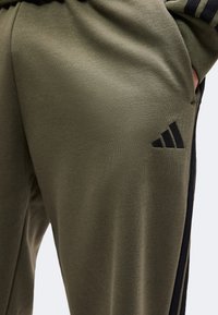 Κυπαρισσί πράσινη φόρμα Adidas με μαύρες ρίγες στο πλάι και λογότυπο Adidas κοντά στην τσέπη, το χέρι μερικώς στην τσέπη.