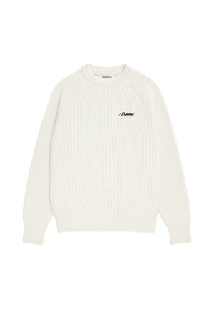 Homme portant un pull côtelé crème avec une écharpe grise à motifs nouée autour du cou et un jean noir délavé, debout devant un fond uni.