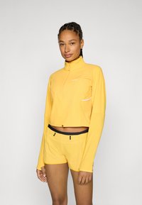 Kollane pika varrukaga zip-up top kõrge kraega ja peegeldavate aktsentidega, millele on sobitatud tekstuurilised kollased lühikesed püksid musta vöökoha ja nööriga.