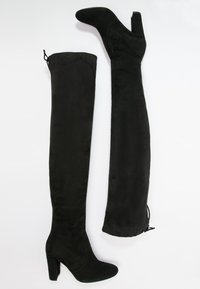 Par de botas negras de ante hasta la rodilla con tacones de bloque, una bota en posición vertical y la otra de lado sobre un fondo blanco.