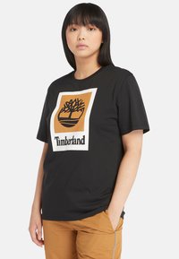 Svart bomull T-skjorte med et sentralt brunt og hvitt Timberland-logo med et tre-grafikk. Korte ermer og rund hals.