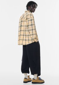 Żółta i zielona koszula w krat�ę, w oversize'owym kroju, w połączeniu z czarnymi spodniami i brązowymi sneakersami z niebieskimi sznurówkami. Gładka faktura i casualowy fason.