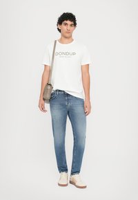 White cotton t-shirt with "DONdup" logo, light blue jeans, and beige sneakers. Model holds a small, gray crossbody bag.