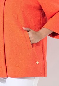Oranžový texturovaný kabát s vzorem listů, předními kapsami a zlatým akcentem u spodního okraje. Bílé kalhoty jsou částečně viditelné.