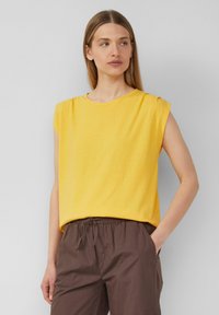 Yellow short-sleeve top made of soft fabric with a loose fit, featuring a round neckline and shoulder pleats. Paired with brown elastic waistband pants.